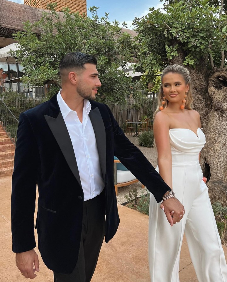 a man in a tuxedo and a woman in a white jumpsuit hold hands