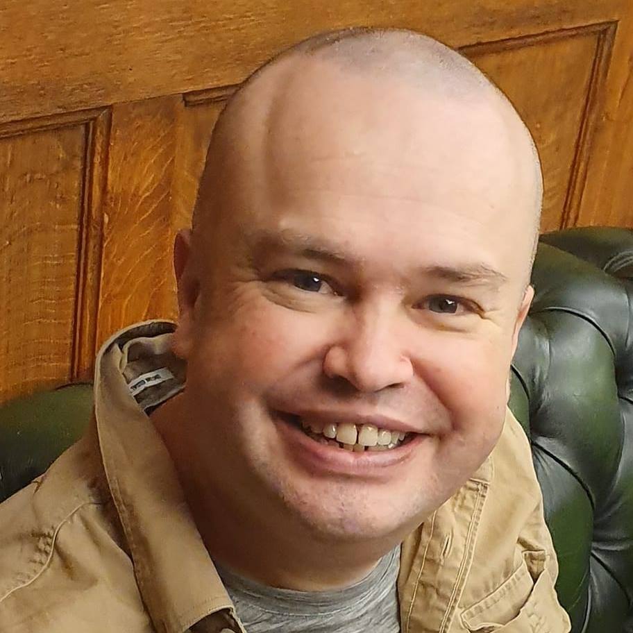 a bald man wearing a tan jacket smiles for the camera