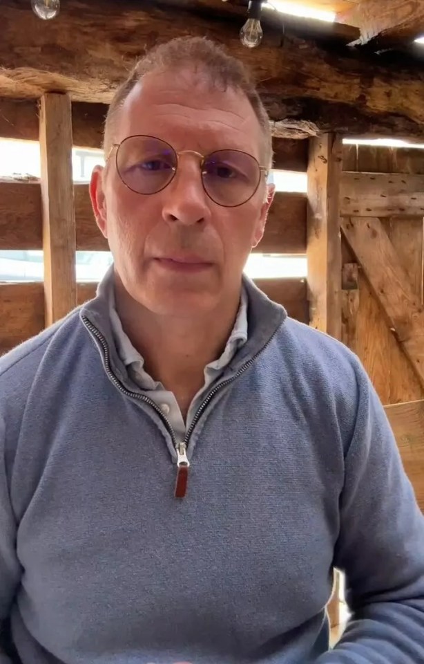 a man wearing glasses and a blue sweater stands in front of a wooden wall