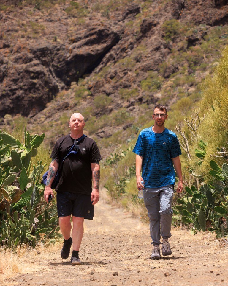 two men are walking down a dirt path with cactus in the background