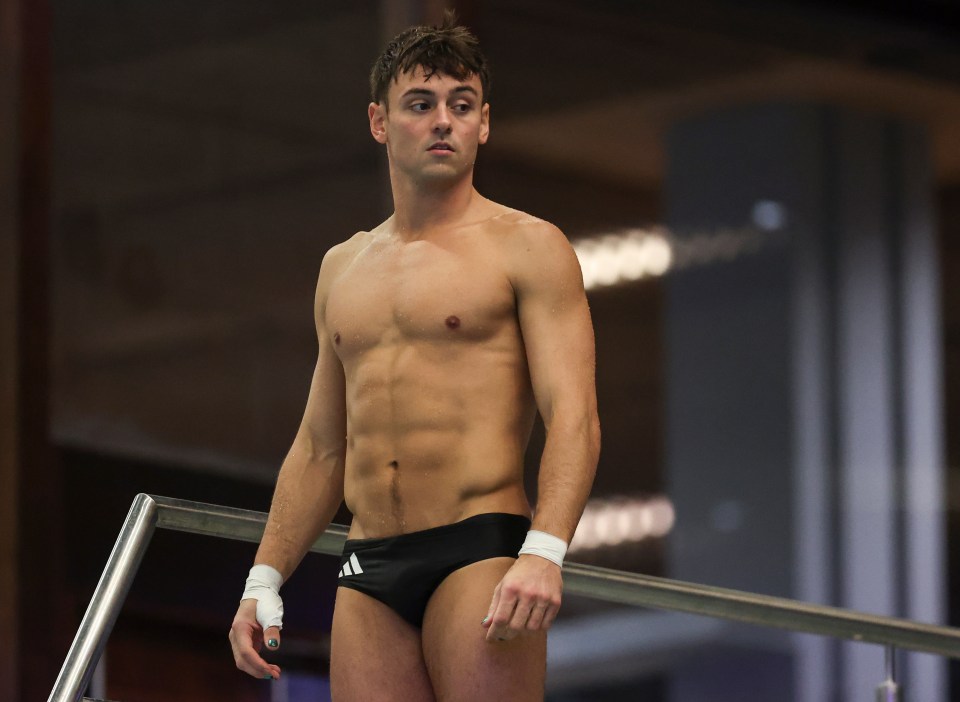 a shirtless man in black adidas swim trunks stands on a diving board