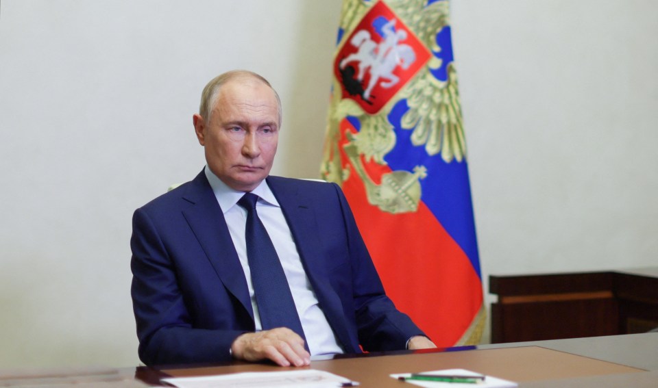 a man in a suit and tie sits at a desk in front of a russian flag