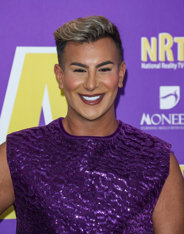 a man in a purple dress smiles in front of a sign that says nri