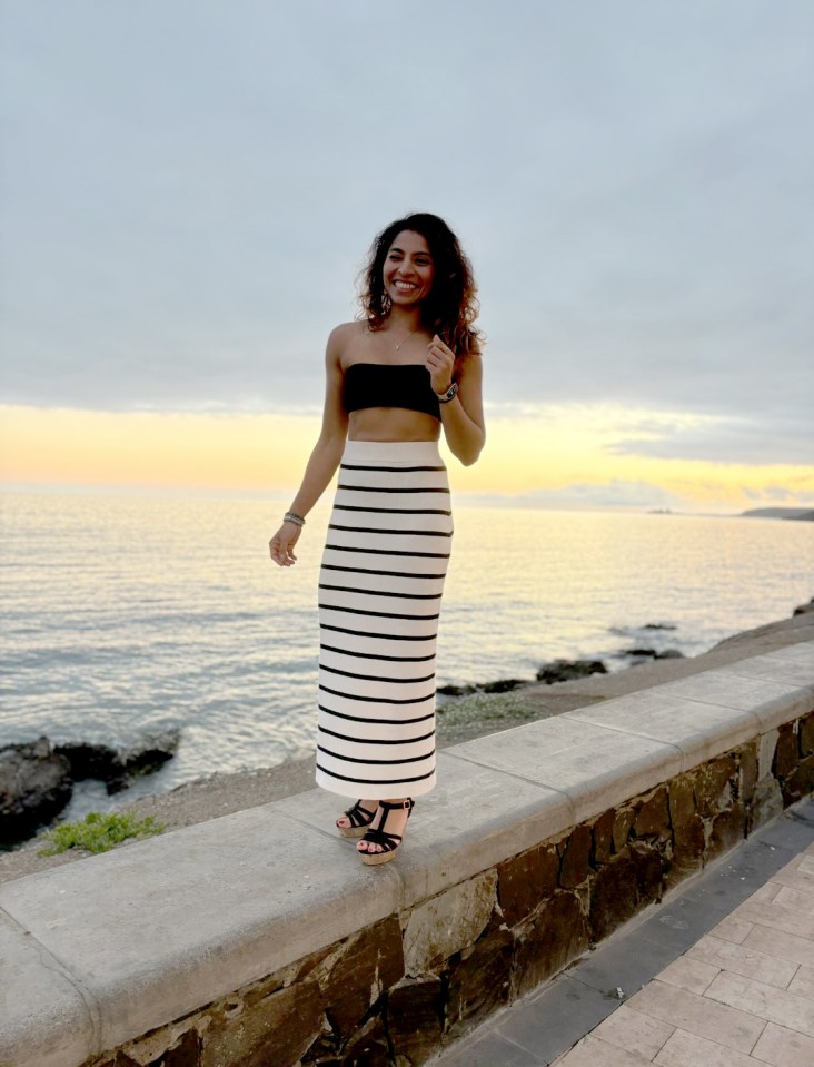 a woman in a black and white striped skirt stands on a stone wall near the ocean