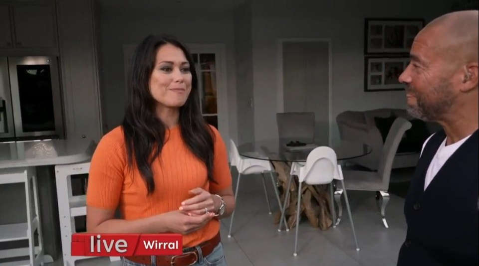 a woman in an orange shirt is standing next to a bald man in a living room with a live wirral sign behind her