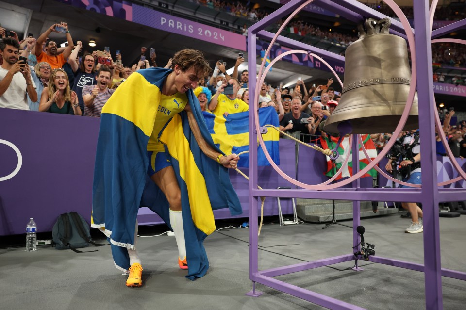 a man wrapped in a swedish flag stands in front of a bell that says paris 2024