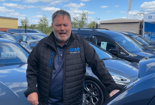 a man wearing a one automotive jacket stands in front of a row of cars