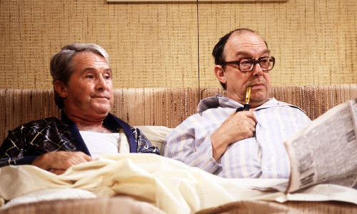 two men are laying on a couch reading newspapers