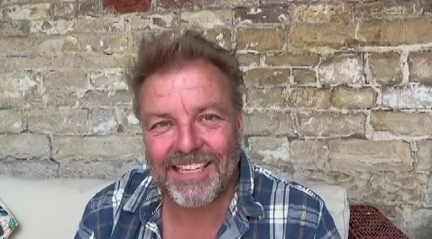 a man with a beard is smiling while sitting on a couch in front of a brick wall .