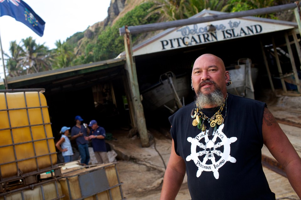 a man wearing a pitcairn island shirt stands in front of a building