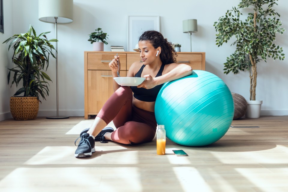 a woman sits on the floor eating a bowl of cereal next to an exercise ball