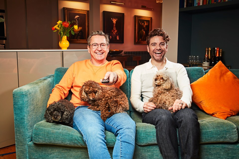 two men are sitting on a couch with their dogs and one is holding a remote control