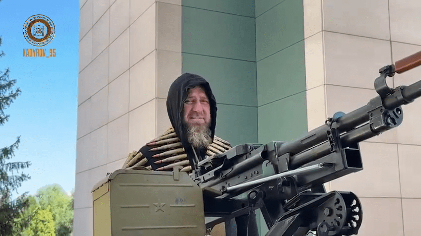 Kadyrov stands on the bed behind the gun with the ammo belt around his shoulders