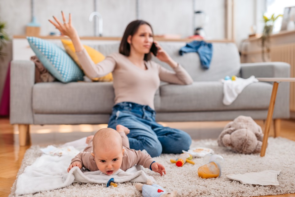 Young tired and sleepy mother sitting next to her crying baby boy on the floor in the living room and talking on the phone.