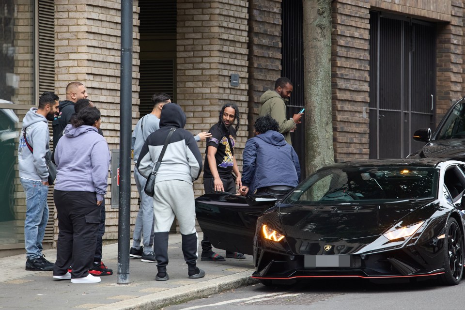 a group of people standing around a black car with a license plate that has the letter w on it