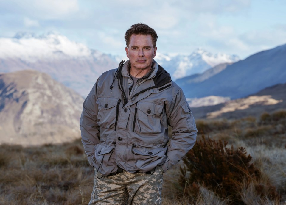 a man in a camouflage jacket stands in front of a mountain