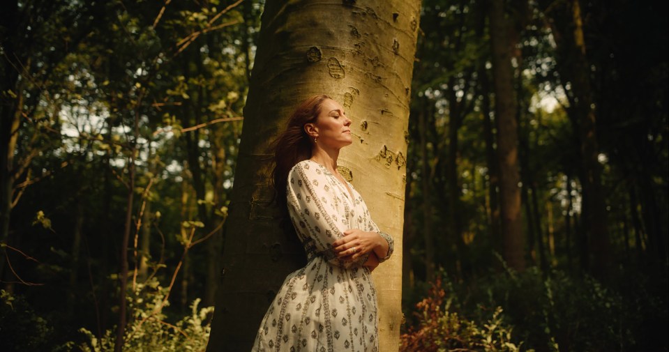 a woman in a white dress leans against a tree in the woods