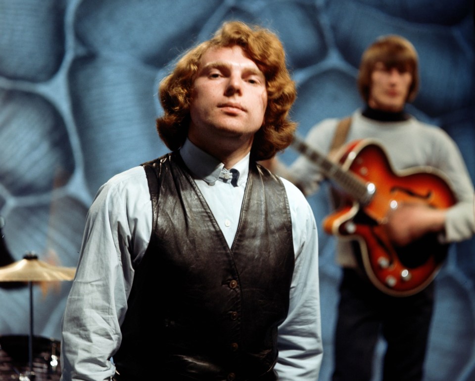 a man in a leather vest stands in front of another man playing a guitar