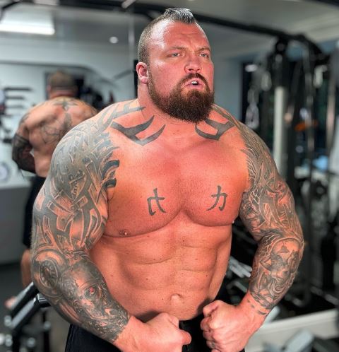 a man with a beard and tattoos is standing in front of a mirror in a gym .