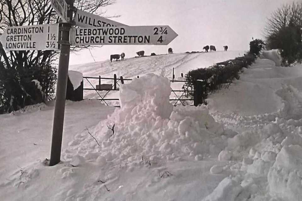 a black and white photo of a snowy field with a sign pointing to leebotwood church stretton