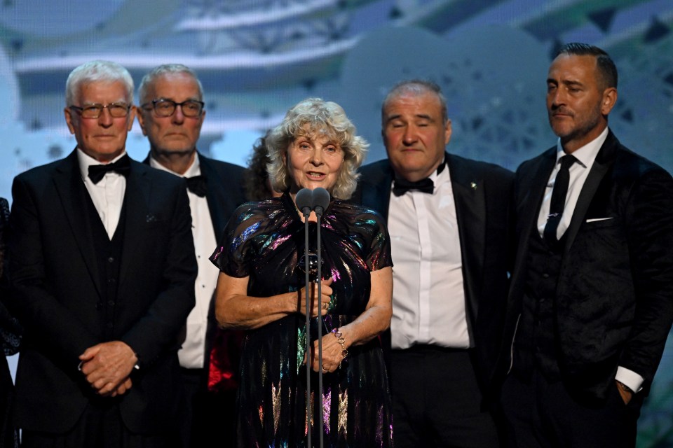 a woman stands in front of a microphone surrounded by men in tuxedos