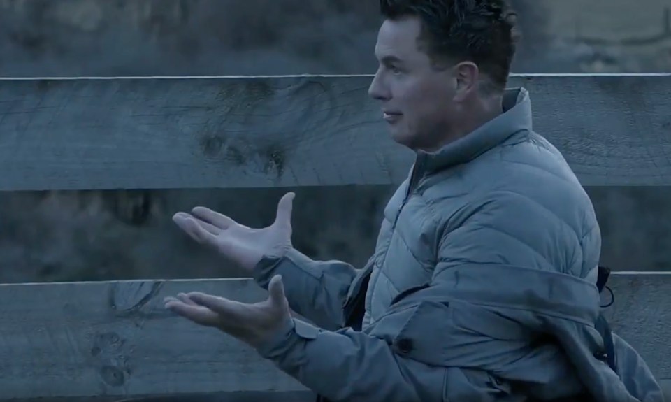 a man in a grey jacket is standing in front of a wooden fence