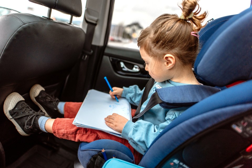 a little girl sits in a car seat drawing on a piece of paper