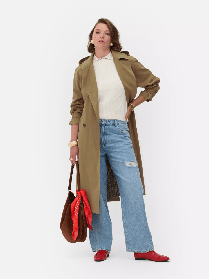 a woman wearing a trench coat and jeans is holding a red bag