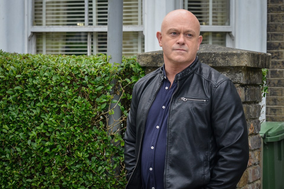 a bald man in a black leather jacket stands in front of a bush