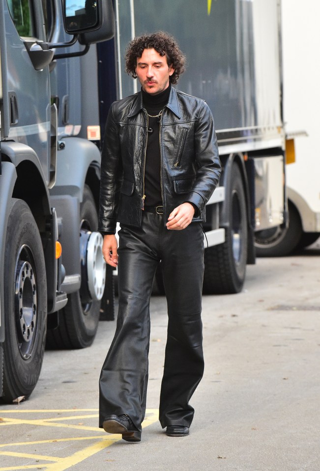 a man wearing a leather jacket and wide leg pants walks down a street