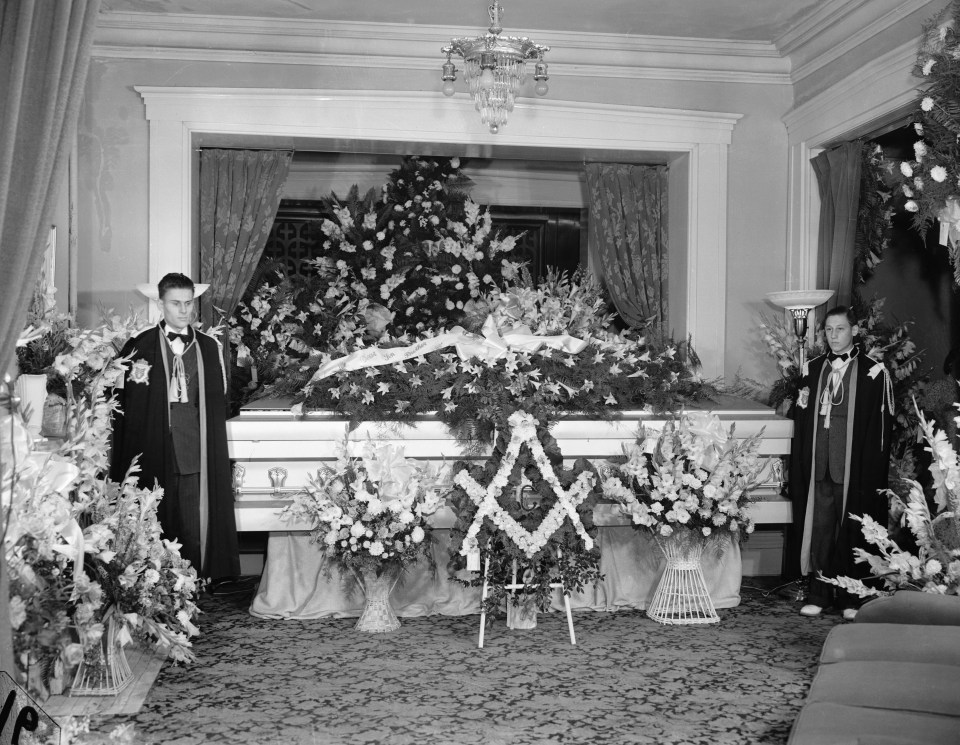 two men stand in front of a white coffin decorated with flowers