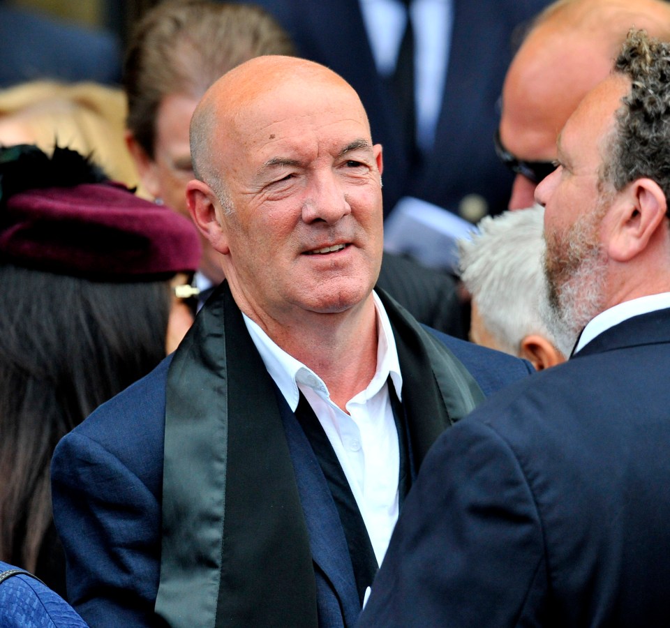 a bald man in a suit talks to another man in a crowd