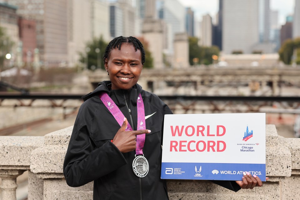 a woman in a black nike jacket smiles in front of a blue wall that says chicago marathon