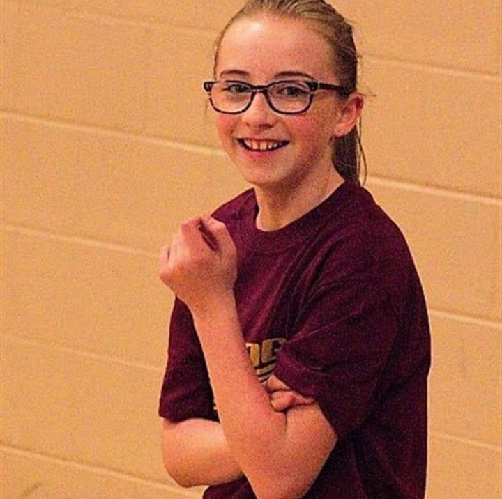 a young girl wearing glasses and a maroon shirt is standing in front of a brick wall .