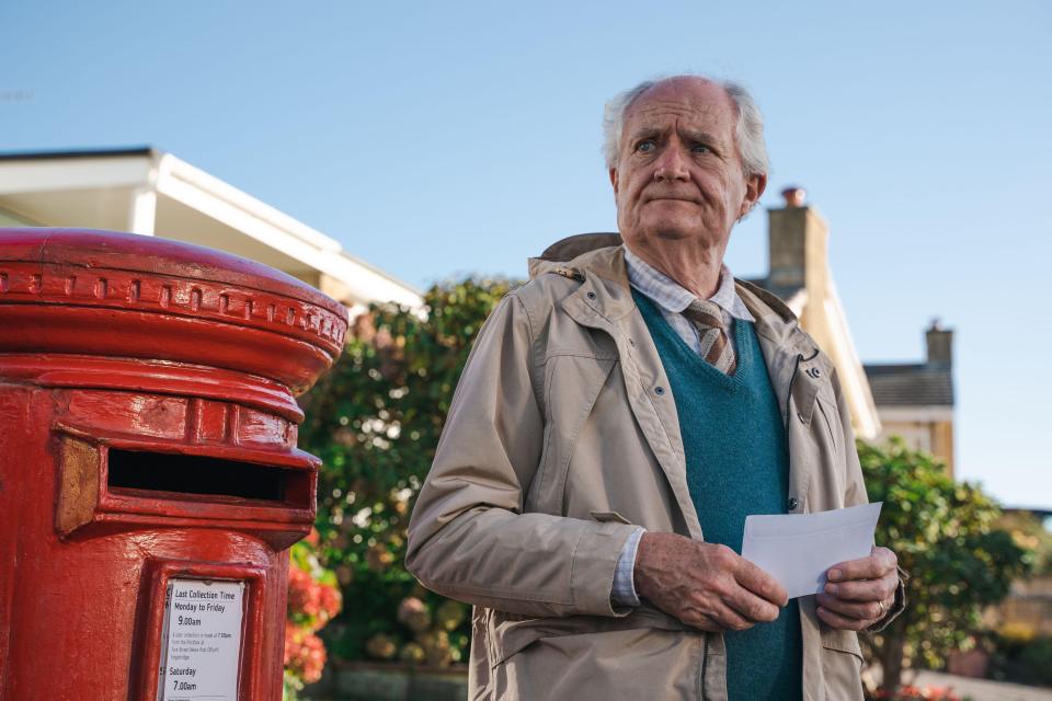 an elderly man is holding a piece of paper in front of a red post box