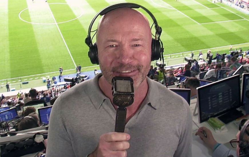 a bald man wearing headphones holds a microphone in front of a soccer field