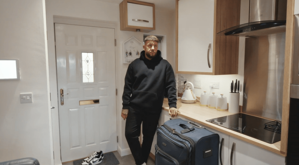 a man in a black hoodie stands in a kitchen with a blue suitcase