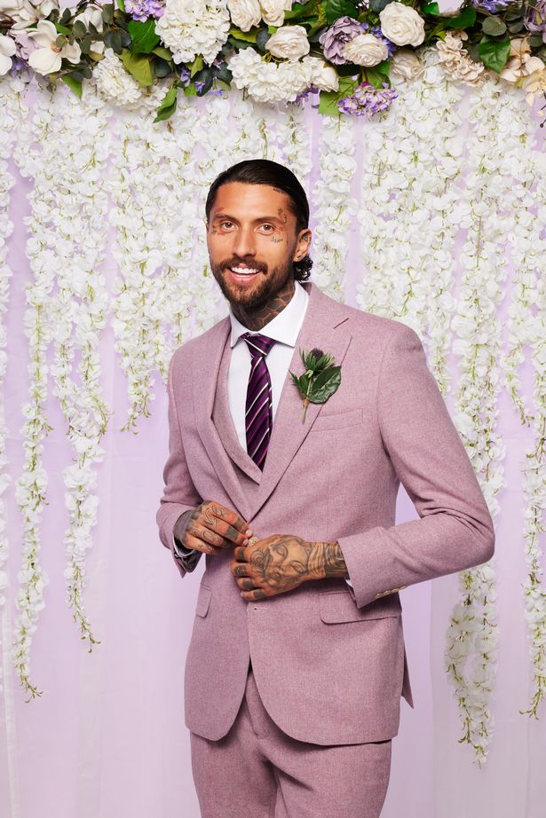 a man in a purple suit and tie stands in front of a wall of white flowers