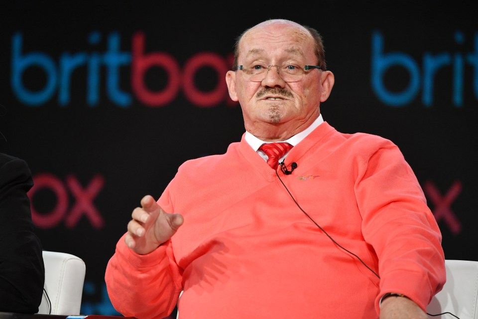 a bald man wearing glasses and a red sweater sits in front of a sign that says britbox