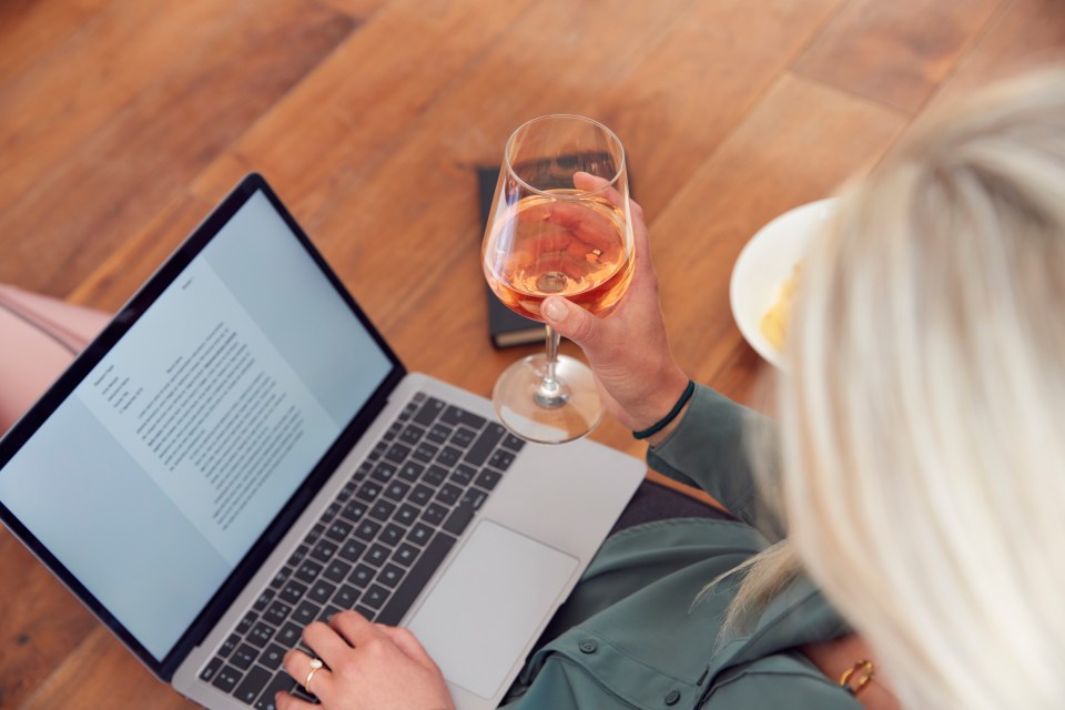 a woman is typing on a laptop while holding a glass of wine