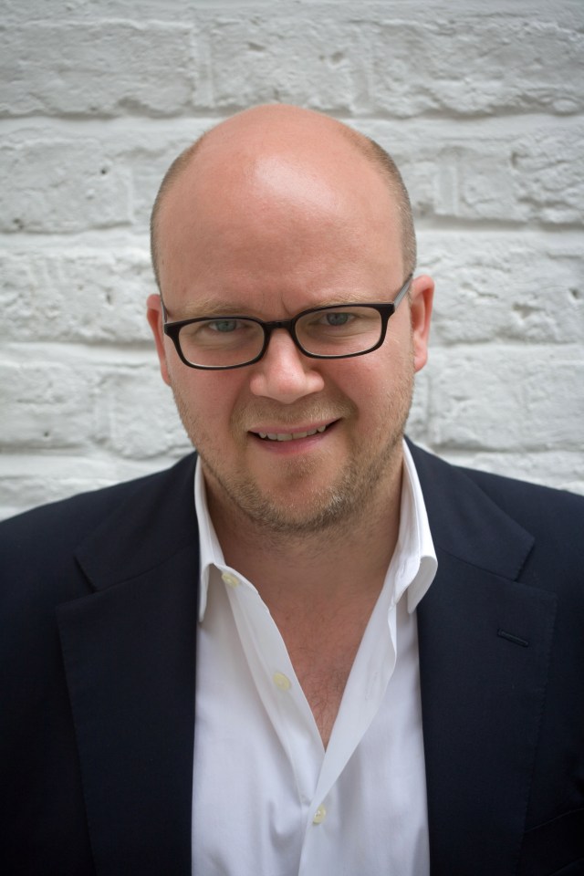 a bald man wearing glasses and a suit smiles in front of a white brick wall