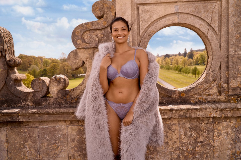 a woman in lingerie and a fur coat stands in front of a stone wall