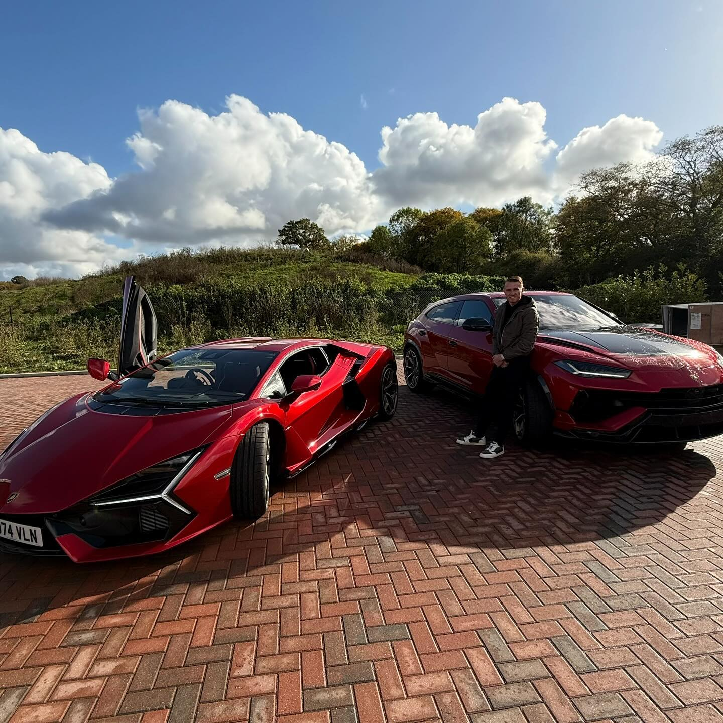 a man stands in front of two red sports cars one of which has a license plate that starts with the letter t