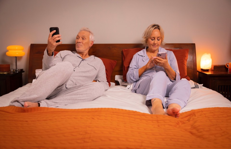 a man and a woman sit on a bed looking at their phones