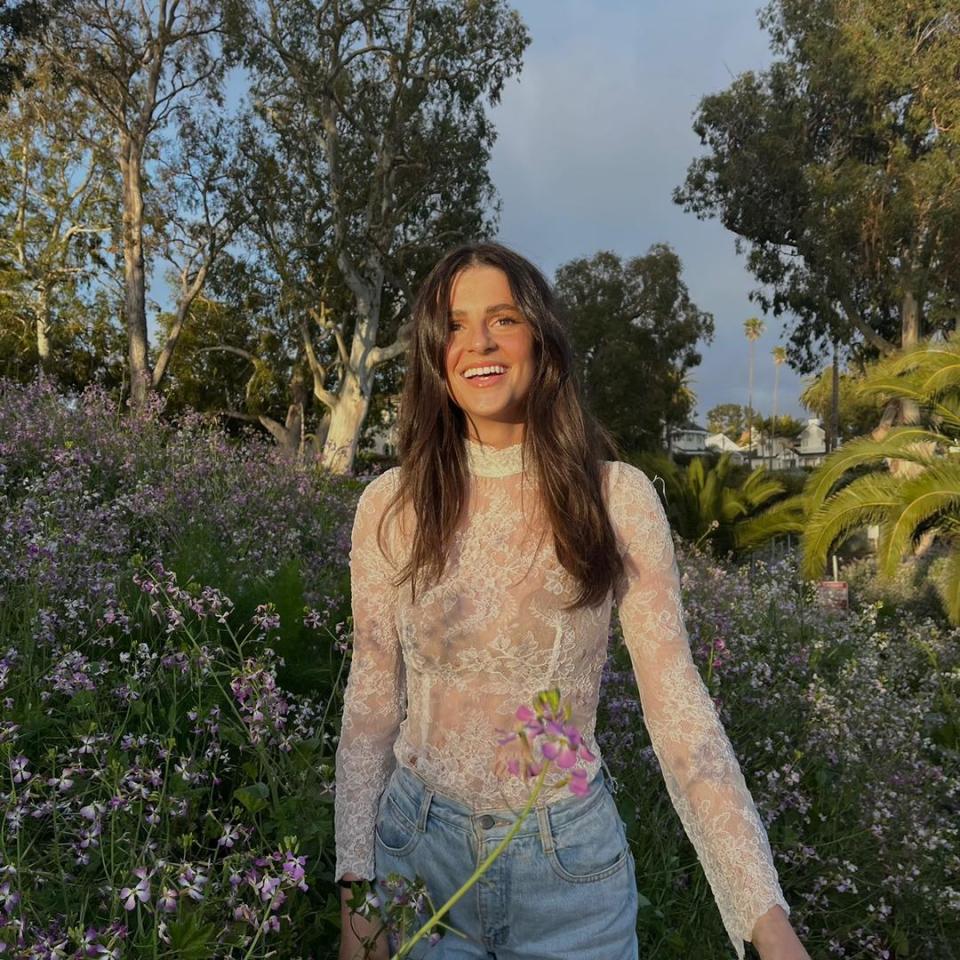 a woman in a white lace top stands in a field of purple flowers