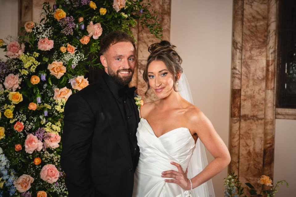 a bride and groom pose in front of a wall of flowers