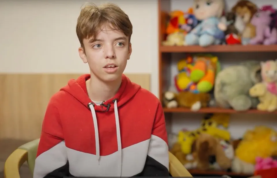 a young boy in a red and white hoodie sits in front of a shelf full of stuffed animals