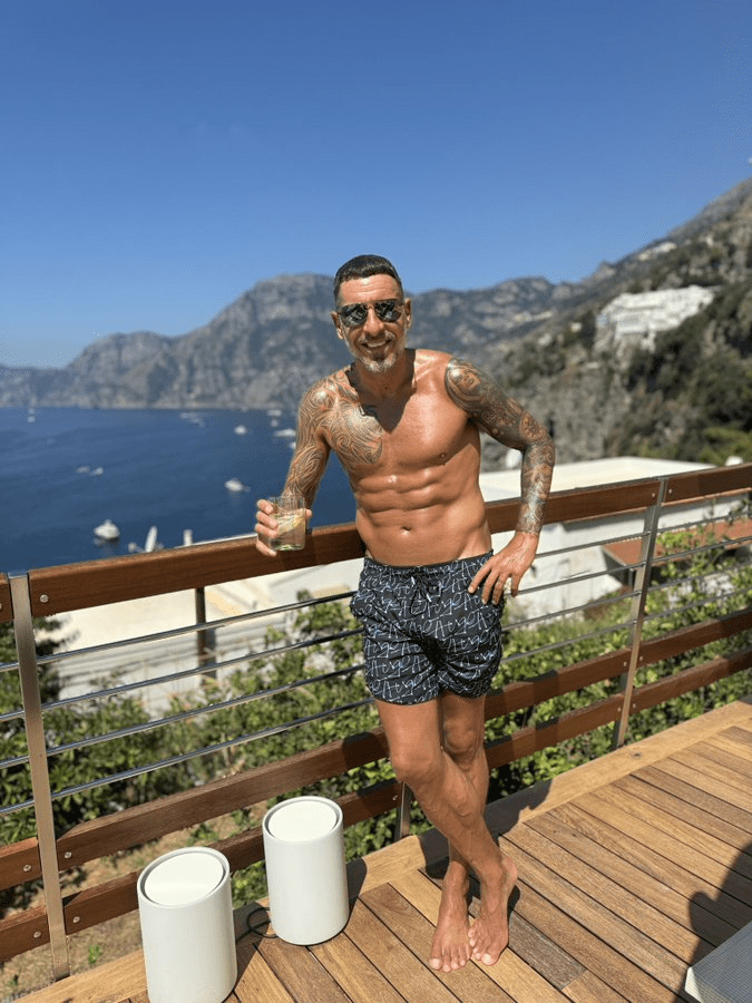 a shirtless man stands on a balcony holding a drink