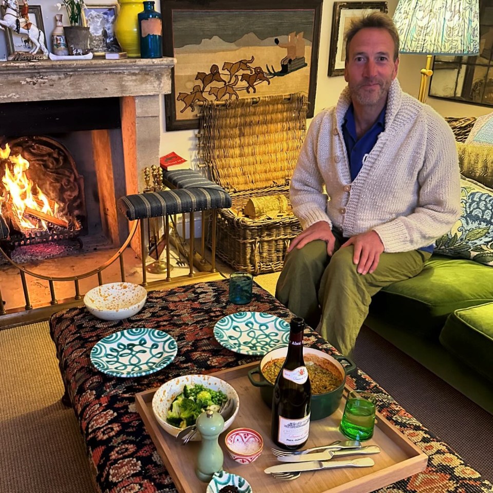 a man sits in front of a fireplace with a tray of food and a bottle of wine