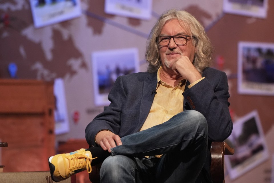 a man with long hair and glasses sits in a chair with his legs crossed